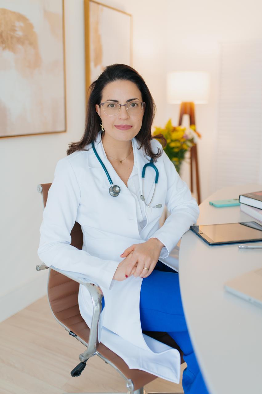 Dra. Gerusa Vilas-Bôas sentada no consultório, de jaleco branco, com o estetoscópio ao pescoço.
